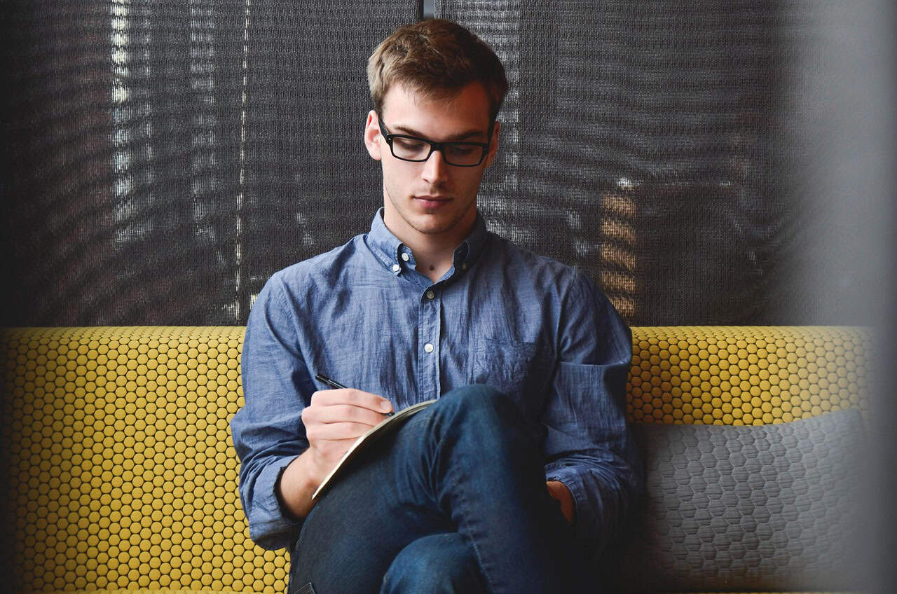Young man writing in a notebook, sitting on a yellow patterned sofa near a window.